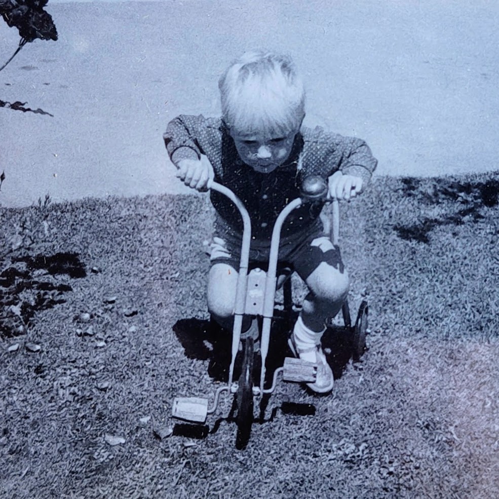 A small blond boy head-down on a tricycle, black and white